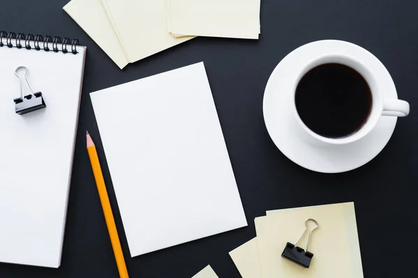 Draufsicht auf leeres Papier, Notizbuch, Bleistift, Klammern und Tasse Kaffee auf Schwarz — Stockfoto