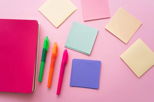 Vista superior de plumas de colores cerca de cuaderno brillante y notas de papel en blanco en rosa - foto de stock