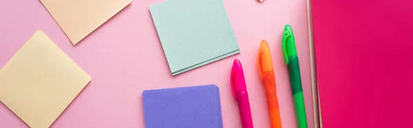 Top view of colorful pens near bright notebook and blank paper notes on pink, banner — Stock Photo