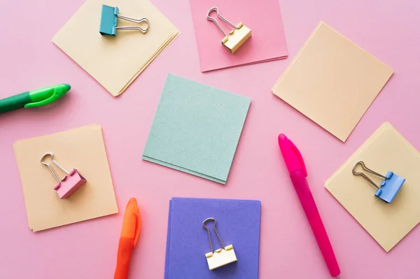 Flat lay of fold back clips on colorful paper notes near pens on pink — стоковое фото