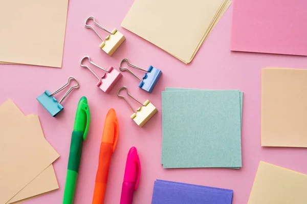 Top view of fold back clips near paper notes and colorful pens on pink — стоковое фото