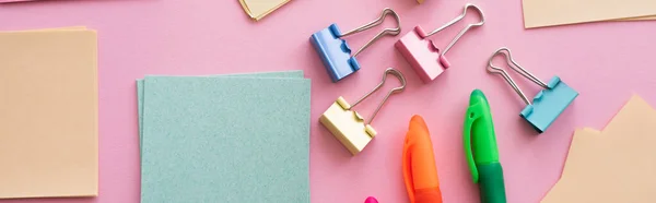 Top view of fold back clips near paper notes and colorful pens on pink — стоковое фото