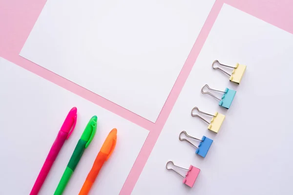 Flat lay of colorful fold back clips and pens on white papers on pink - foto de stock