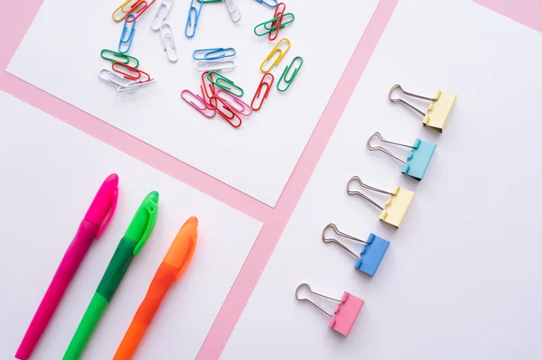 Flat lay of colorful fold back clips, paper clips and pens on white papers on pink - foto de stock