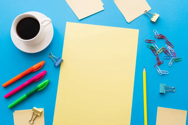 Vue du dessus de tasse de café et de papiers vides près de stylos colorés et trombones sur bleu — Photo de stock