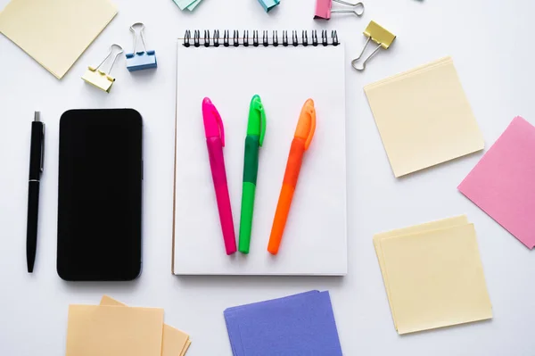 Top view of smartphone with blank screen near notebook with colorful pens and stationery on white — стоковое фото