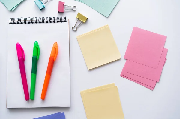 Vista dall'alto di penne colorate su notebook vicino a note di carta vuote e piegare indietro clip su bianco — Foto stock