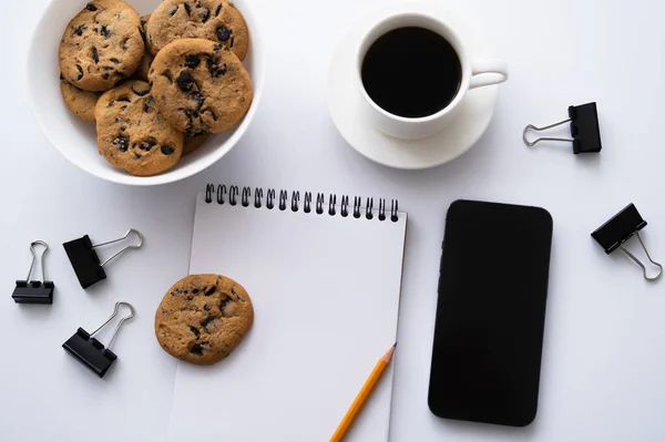 Vista superior de café, biscoitos e artigos de papelaria perto do smartphone com tela em branco no branco — Fotografia de Stock