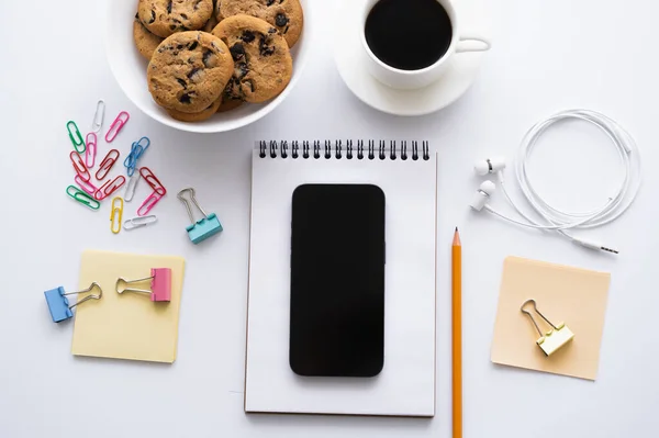 Top view of cup of coffee, cookies and smartphone with blank screen near stationery on white — стоковое фото