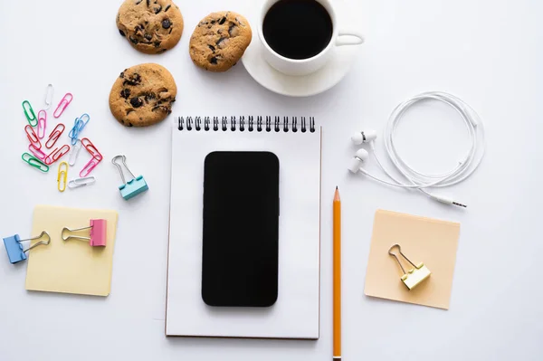 Vista superior da xícara de café, biscoitos e smartphone com tela em branco perto de artigos de papelaria em branco — Fotografia de Stock