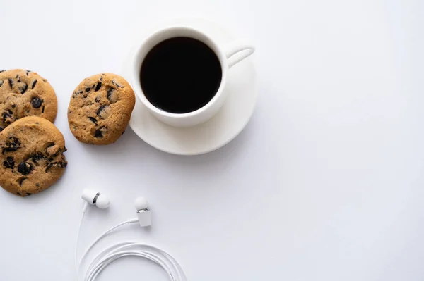 Vue du dessus de tasse de café près de biscuits aux pépites de chocolat et écouteurs filaires sur blanc — Photo de stock