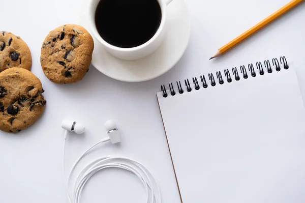 Vue du dessus de tasse de café près de biscuits aux pépites de chocolat, papeterie et écouteurs filaires sur blanc — Photo de stock