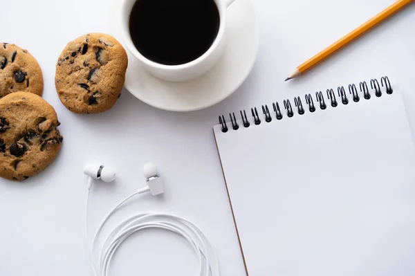 Vista superior da xícara de café perto de biscoitos de chocolate, artigos de papelaria e fones de ouvido com fio no branco — Fotografia de Stock
