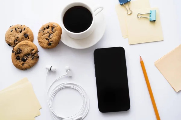 Vue du dessus de tasse de café et de biscuits aux pépites de chocolat près de la papeterie et smartphone sur blanc — Photo de stock