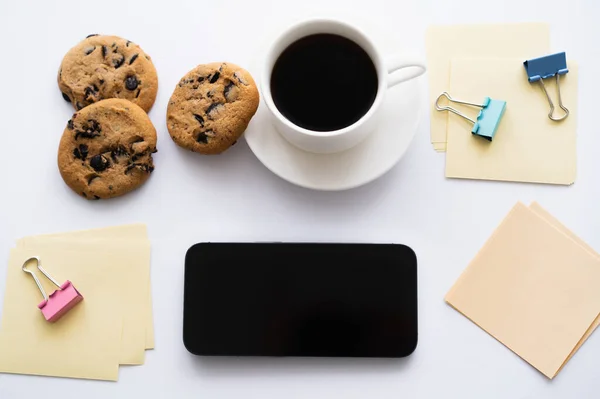 Top view of cup of coffee and chocolate chip biscuits near stationery and smartphone on white — Foto stock