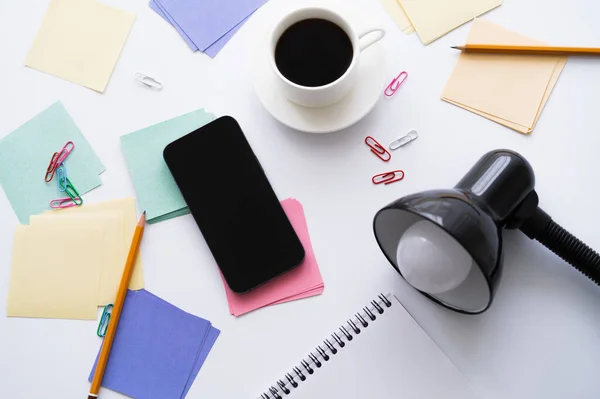 Top view of cup of coffee near stationery, lamp and smartphone with blank screen on white — Photo de stock