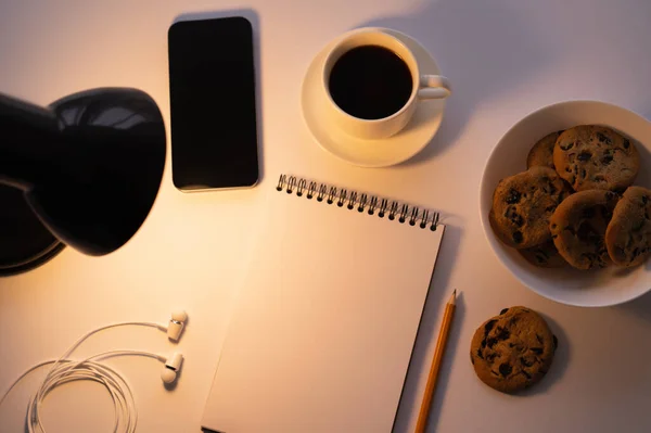 Вид зверху на лампу біля смартфона, печиво з шоколадними чіпсами, чашка кави та блокнот на білому — стокове фото