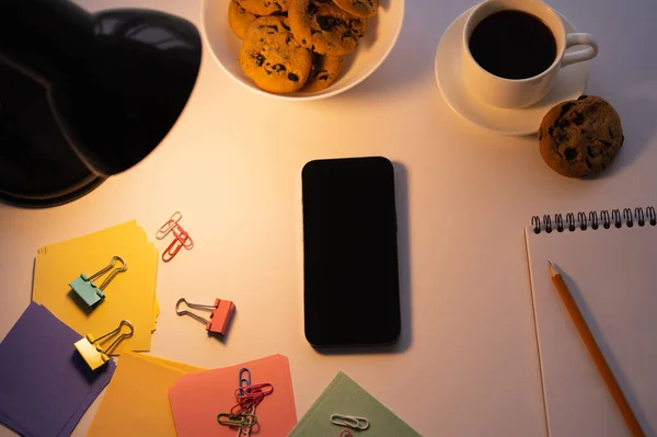 Vue du dessus de la lampe près du smartphone avec écran blanc, biscuits aux pépites de chocolat, tasse de café et papeterie sur blanc — Photo de stock