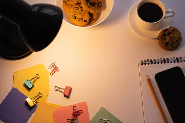 Vista superior da lâmpada perto do smartphone com tela em branco, cookies, xícara de café e artigos de papelaria em branco — Fotografia de Stock