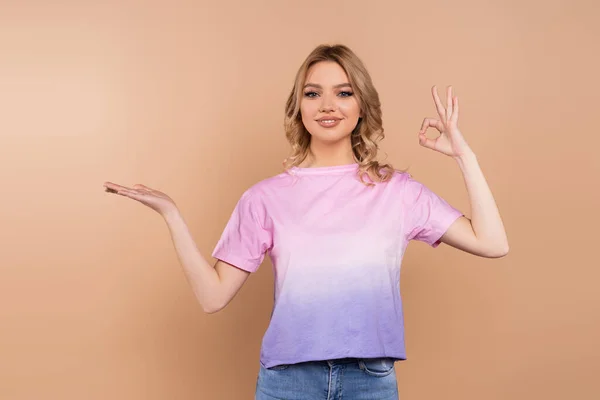 Happy woman pointing with hand and showing okay sign isolated on beige — Stock Photo
