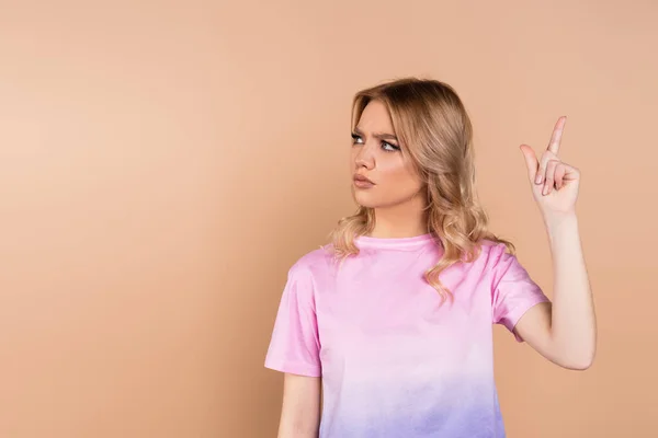 Frowning woman looking away and pointing up with finger isolated on beige — Stock Photo