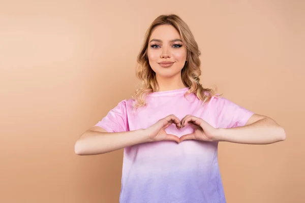 Mujer positiva con el pelo ondulado que muestra el símbolo del corazón con las manos aisladas en beige - foto de stock