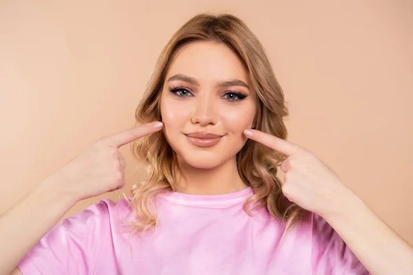 Cheerful woman pointing with fingers at her smile isolated on beige — Stockfoto