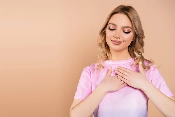 Femme heureuse et reconnaissante avec les cheveux ondulés toucher poitrine isolé sur beige — Photo de stock