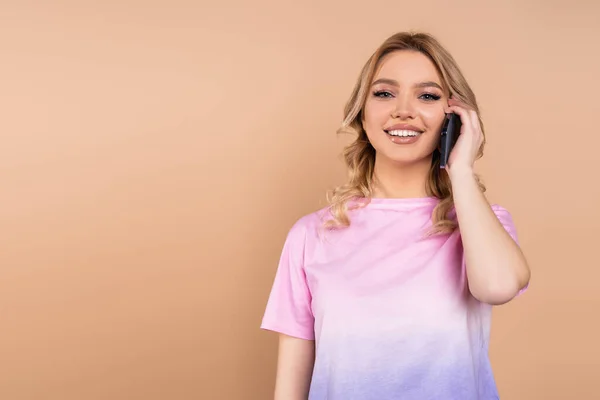 Joyful woman in t-shirt talking on smartphone while looking at camera isolated on beige — Stockfoto