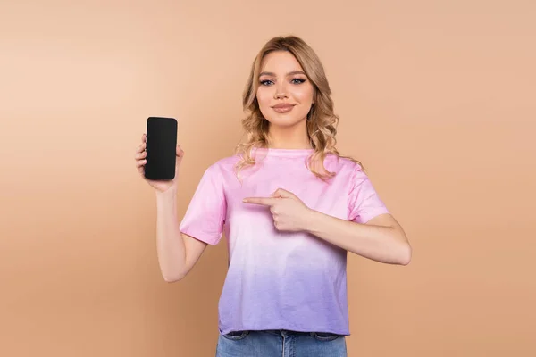 Smiling woman pointing with finger at smartphone with blank screen isolated on beige — стоковое фото
