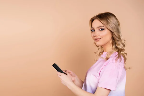 Zufriedene Frau blickt in Kamera, während sie Handy isoliert auf Beige benutzt — Stockfoto