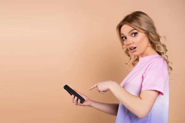 Amazed woman pointing at mobile phone and looking at camera isolated on beige — Stockfoto
