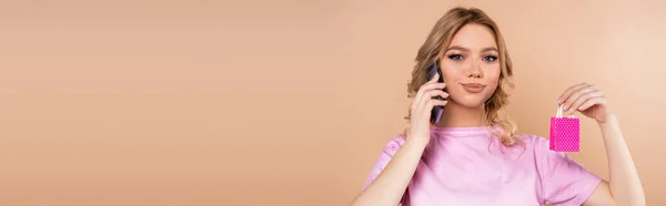 Smiling woman with tiny shopping bag talking on smartphone isolated on beige, banner — Stockfoto