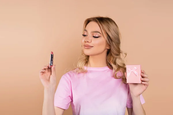 Pleased woman holding lipstick and small gift box isolated on beige — Stock Photo