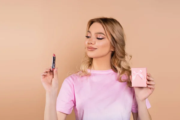 Pleased woman holding lipstick and small gift box isolated on beige — Fotografia de Stock