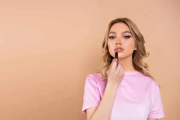 Young woman looking at camera while applying lipstick isolated on beige — стоковое фото