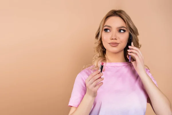 Happy woman talking on mobile phone while holding lipstick isolated on beige — стоковое фото