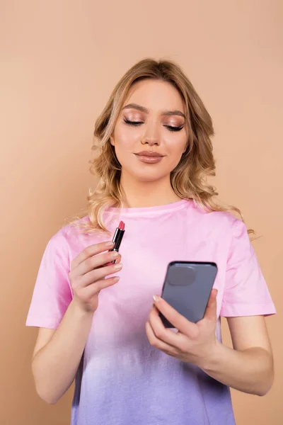 Jolie femme tenant rouge à lèvres et regardant téléphone mobile isolé sur beige — Photo de stock