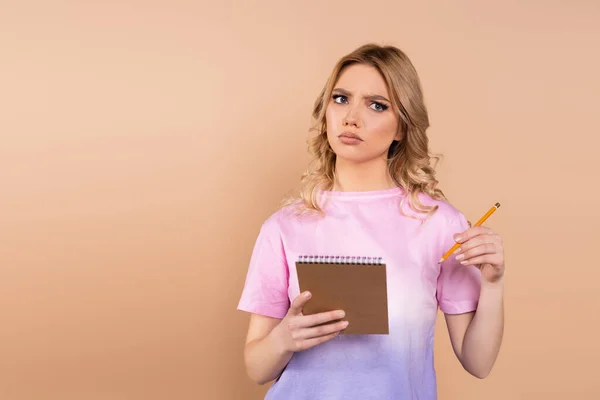 Mujer pensativa con lápiz y bloc de notas mirando hacia otro lado aislado en beige - foto de stock