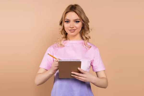 Pretty woman smiling at camera while writing in notebook isolated on beige — Stock Photo