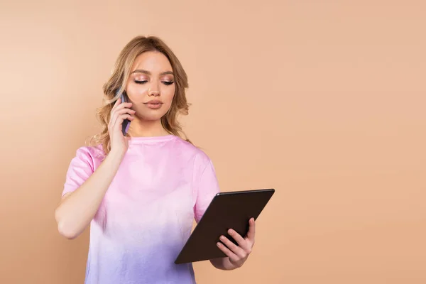 Junge Frau spricht auf Smartphone und blickt vereinzelt auf digitales Tablet in Beige — Stockfoto