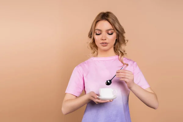 Young woman with wavy hair holding coffee cup and teaspoon isolated on beige — Photo de stock