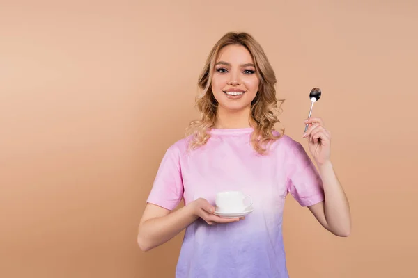 Mujer alegre con taza de café y cucharadita mirando a la cámara aislada en beige - foto de stock