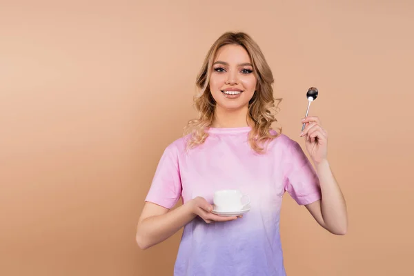 Cheerful woman with coffee cup and teaspoon looking at camera isolated on beige - foto de stock