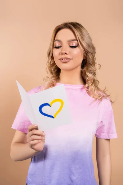 Zufriedene junge Frau liest Karte mit blauem und gelbem Herz isoliert auf beige — Stockfoto