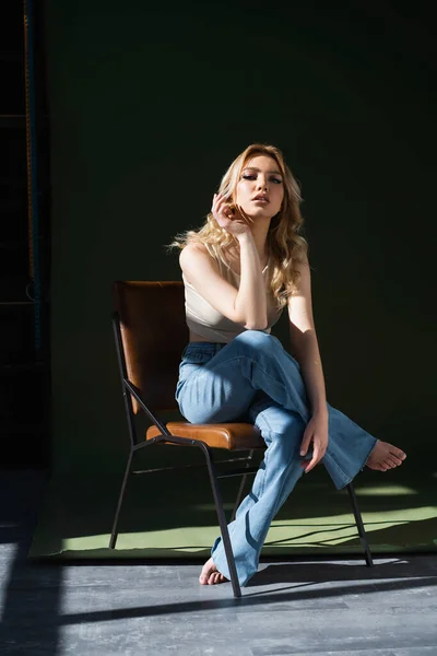 Young barefoot woman in jeans sitting on chair with crossed legs on dark background — Stock Photo