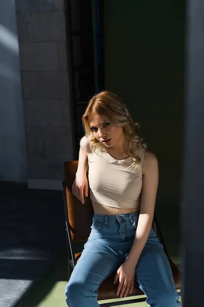Sensual woman in beige top and jeans sitting on chair and looking at camera on blurred foreground - foto de stock