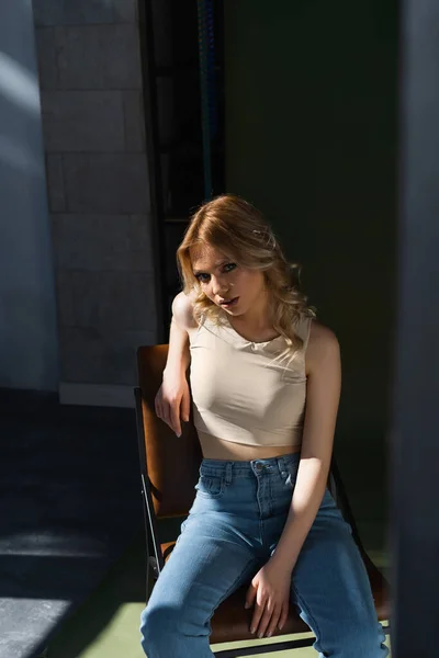 Sensual woman in beige top and jeans sitting on chair and looking at camera on blurred foreground — Fotografia de Stock