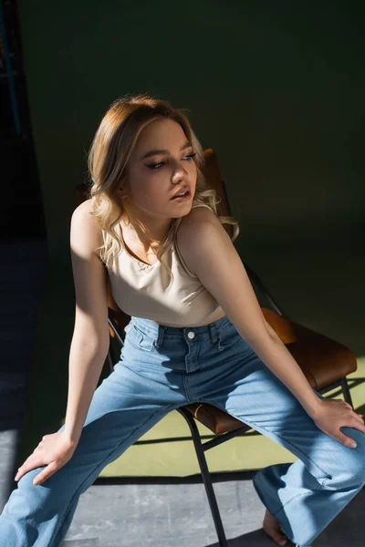 Pretty woman in top and jeans sitting on chair and looking away on dark background — Foto stock