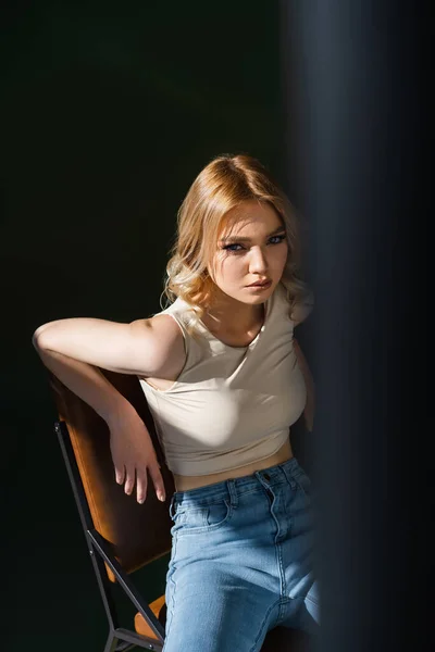 Young woman with wavy hair sitting on chair and looking at camera on black background - foto de stock